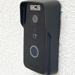 Czarny wideodomofon BCS zamontowany na białej, chropowatej ścianie zewnętrznej, z widocznym przyciskiem dzwonka i logo producenta.