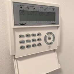 Centrala alarmowa Satel Integra z otwartą klapką, wyświetlająca datę 28 Luty i godzinę 09:20:13 na białej ścianie.