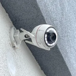 Biała kamera monitoringu Ezviz z antenami zamontowana na szarej, chropowatej ścianie pod wystającym elementem architektonicznym.