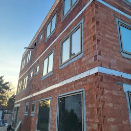 COMFY Piotr Puszkarski - Budynek w trakcie budowy z zamontowanymi oknami w surowym stanie, widoczne bloczki ceramiczne i zaprawa, częściowo otynkowane nadproża, widok z dołu na narożnik budynku.