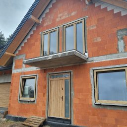 COMFY Piotr Puszkarski - Nowy dom w trakcie budowy, widoczne zamontowane okna z jasną ramą, drzwi wejściowe z przeszkleniem oraz brama garażowa, ściany z czerwonej cegły.