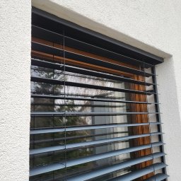 COMFY Piotr Puszkarski - Szare żaluzje fasadowe zamontowane na oknie z drewnianą ramą, widok z zewnątrz na elewację budynku z fakturą baranka.