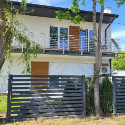 Dwukondygnacyjny dom z balkonem z metalową balustradą, widok zza grafitowego ogrodzenia panelowego, drzewa po bokach.