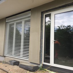 COMFY Piotr Puszkarski - Białe okno z szybą refleksyjną i białe żaluzje poziome na elewacji budynku w trakcie wykończenia, widoczne przewody i gruz na ziemi.