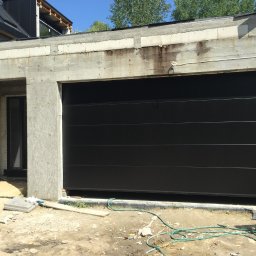 COMFY Piotr Puszkarski - Czarna, segmentowa brama garażowa w surowym otoczeniu budowy, widoczne niedokończone ściany i gruz.