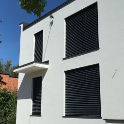 COMFY Piotr Puszkarski - Nowoczesny dom z białymi ścianami i ciemnymi żaluzjami fasadowymi, widok z dołu w słoneczny dzień, z fragmentem zieleni w górnym lewym rogu.