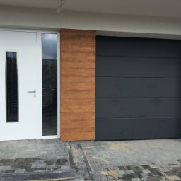 COMFY Piotr Puszkarski - Nowoczesna fasada domu z antracytową bramą garażową segmentową, białymi drzwiami wejściowymi z przeszkleniem i drewnopodobnym panelem dekoracyjnym