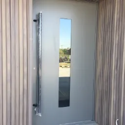 Nowoczesne szare drzwi wejściowe z wąskim przeszkleniem i długim uchwytem zabezpieczonym folią, wkomponowane w elewację z drewnianych paneli.