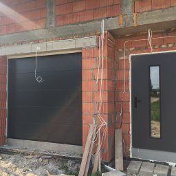 COMFY Piotr Puszkarski - Surowy stan budynku z zamontowaną segmentową bramą garażową w kolorze antracytowym i drzwiami wejściowymi z małym przeszkleniem, widoczne przewody elektryczne i niedokończone mury.