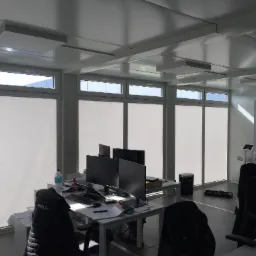 Jasne biuro z rzędem okien z białymi roletami, biurkami z komputerami i krzesłami biurowymi, widoczne oświetlenie sufitowe.