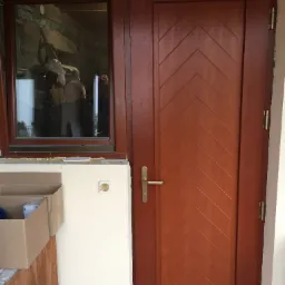 Brązowe drzwi wejściowe z dekoracyjnym wzorem w jodełkę i dopasowane okno boczne, widoczne ślady prac wykończeniowych, kartonowe pudło w pobliżu.