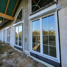 Białe okna z roletami zewnętrznymi w trakcie montażu w budynku z szarej cegły, widoczne materiały izolacyjne i logo firmy na rolecie.