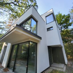 COMFY Piotr Puszkarski - Nowoczesny dom jednorodzinny z dużymi przeszkleniami w czarnych ramach, widok z dołu, otoczony drzewami.