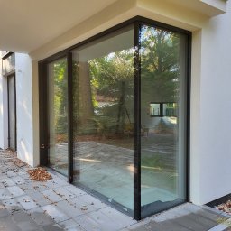 COMFY Piotr Puszkarski - Nowoczesne, panoramiczne przeszklenie z czarnymi ramami w minimalistycznym budynku, widok na ogród, jesienne liście na tarasie z szarych płyt.