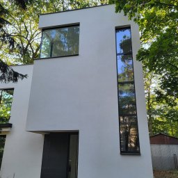 COMFY Piotr Puszkarski - Nowoczesny dom z białymi ścianami, czarnymi ramami okiennymi i wysokim, wąskim oknem, otoczony zielenią drzew.