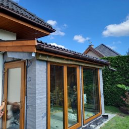 COMFY Piotr Puszkarski - Montaż drewnianej konstrukcji z przeszkleniami, widoczne detale izolacji i folii paroizolacyjnej, pracownik wewnątrz budynku, błękitne niebo w tle.