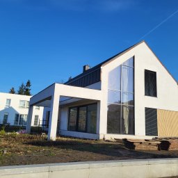 COMFY Piotr Puszkarski - Nowoczesny dom jednorodzinny z dużymi przeszkleniami i żaluzjami zewnętrznymi, widok z zewnątrz, w tle błękitne niebo.