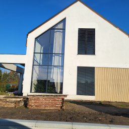 COMFY Piotr Puszkarski - Nowoczesny dom z dużym trójkątnym oknem i ciemnymi żaluzjami poziomymi, widoczne drewniane panele elewacyjne i stosy desek na placu budowy