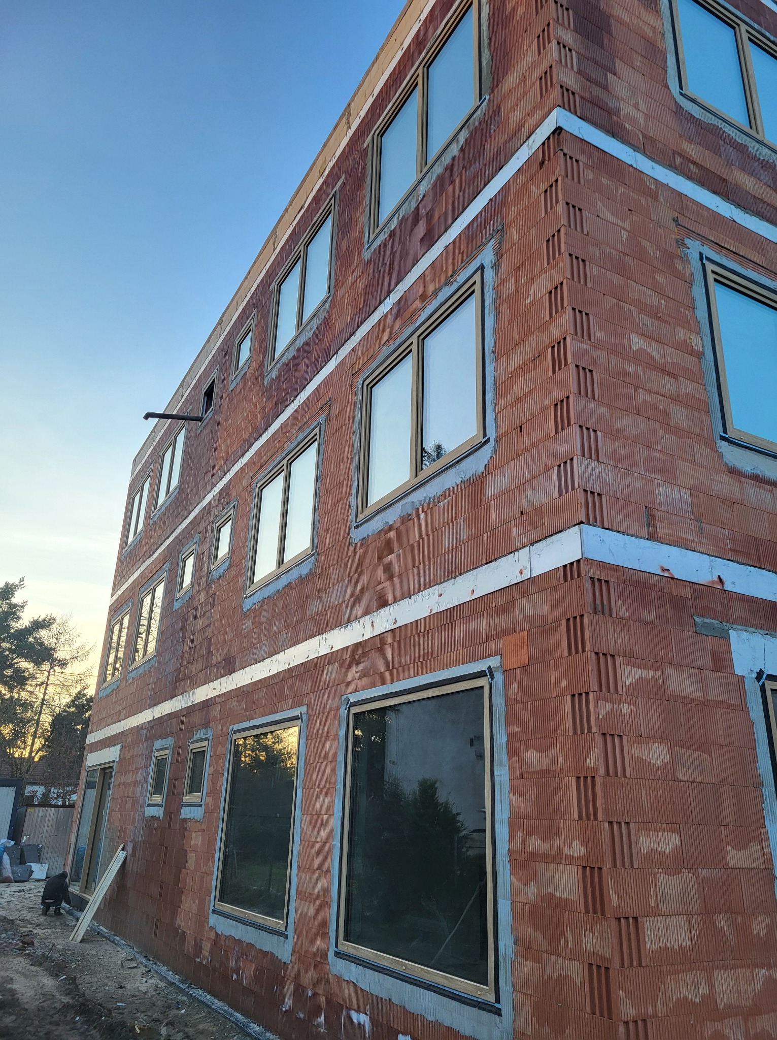 Budynek w trakcie budowy z zamontowanymi oknami w surowym stanie, widoczne bloczki ceramiczne i zaprawa, częściowo otynkowane nadproża, widok z dołu na narożnik budynku.