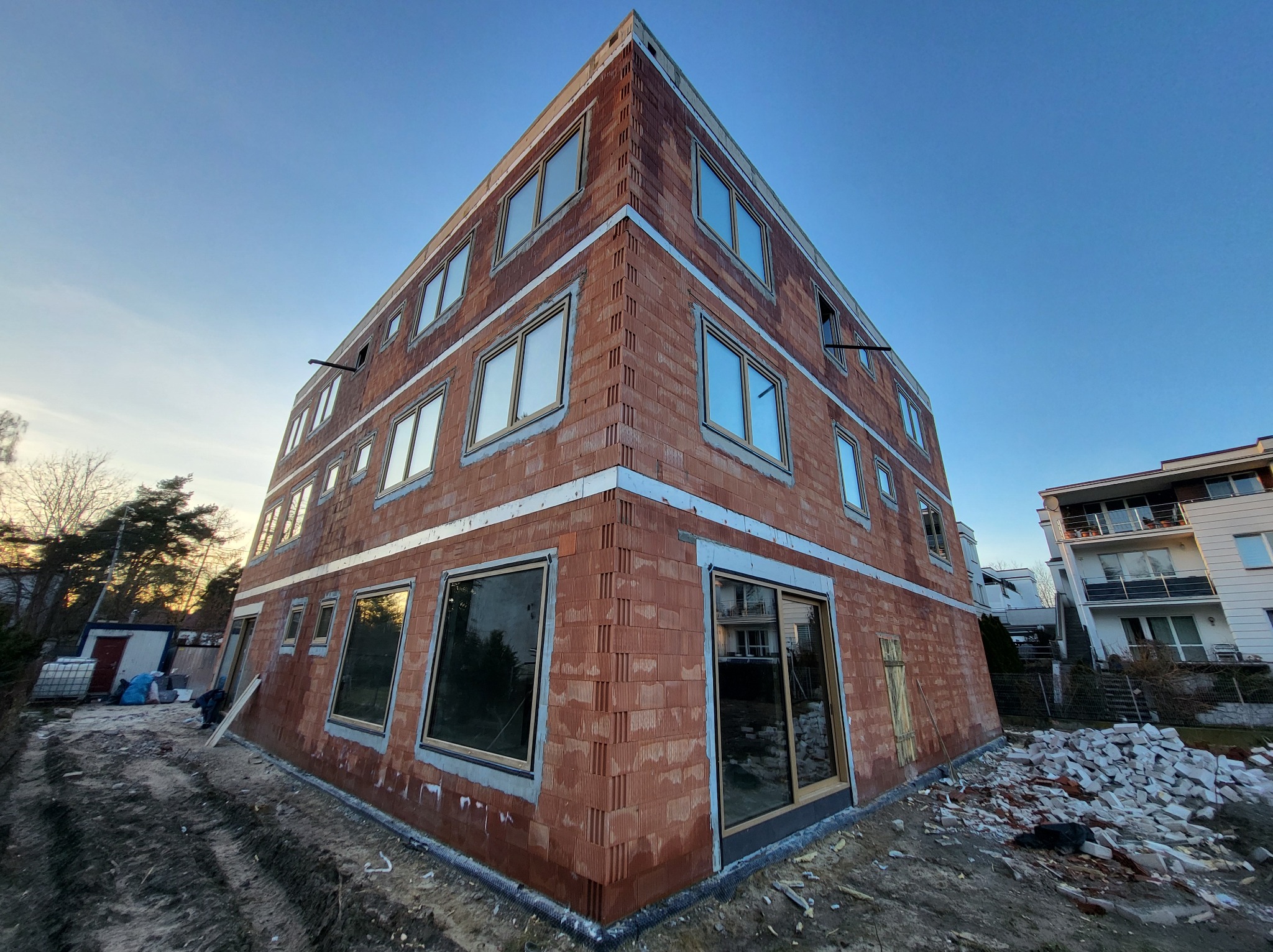 Budynek w trakcie budowy z zamontowanymi oknami, widoczne pustaki ceramiczne i przygotowane otwory okienne, teren budowy z gruzem.