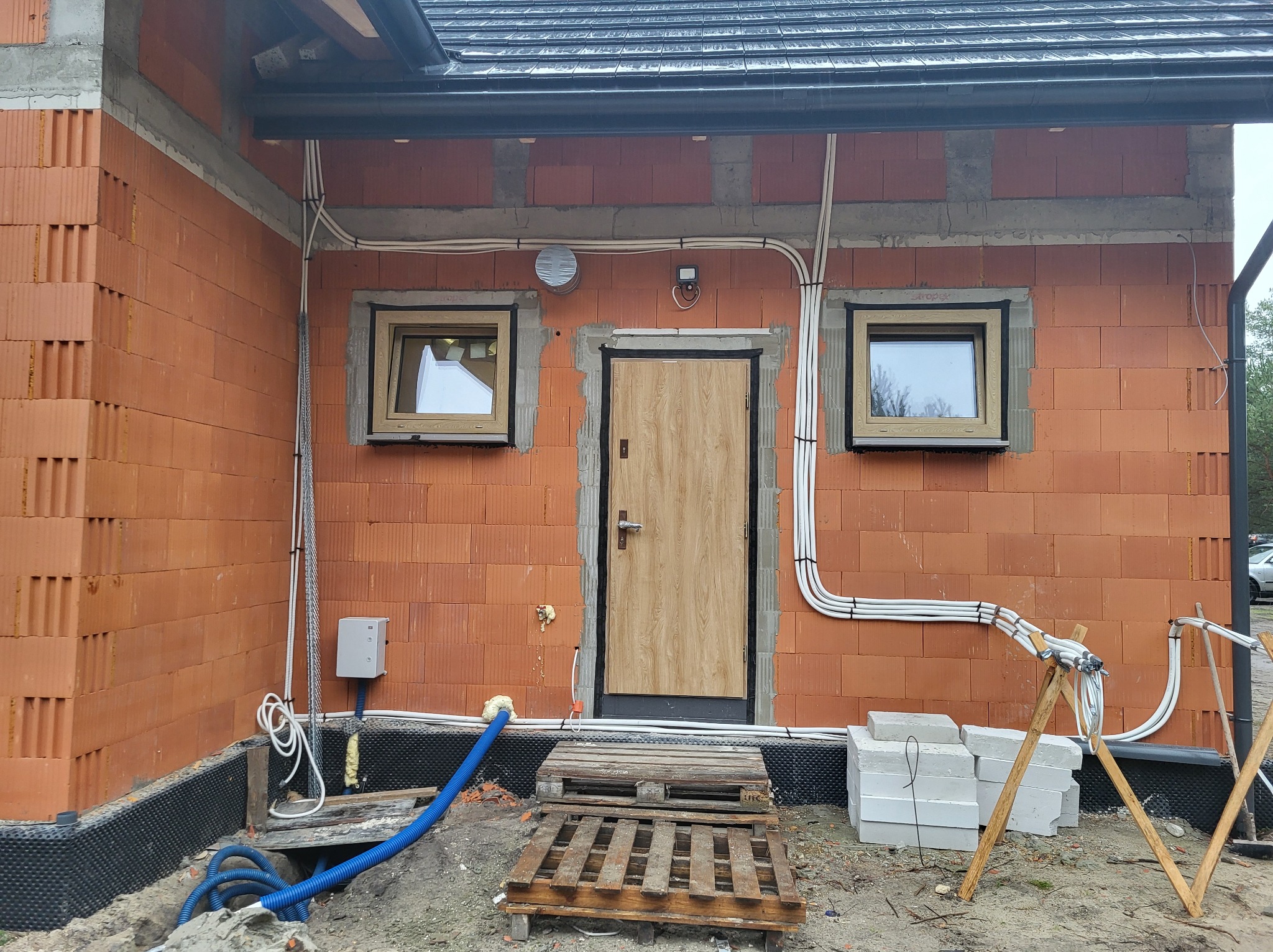 Surowy stan budynku w trakcie wykończenia: zamontowane okna z jasnymi ramami i drzwi wejściowe o fakturze drewna na tle ceglanej ściany, widoczne instalacje elektryczne i elementy izolacji...