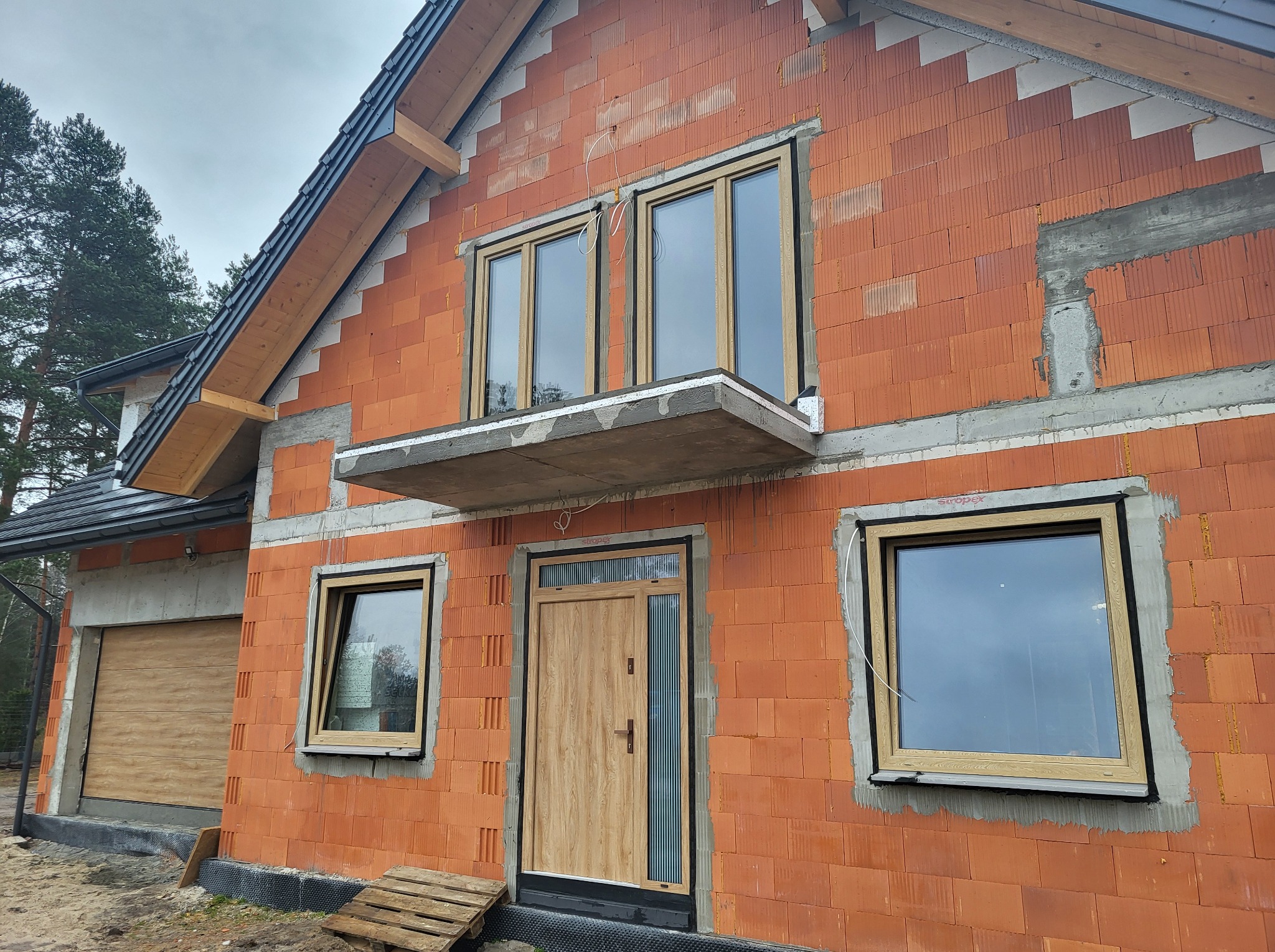 Nowy dom w trakcie budowy, widoczne zamontowane okna z jasną ramą, drzwi wejściowe z przeszkleniem oraz brama garażowa, ściany z czerwonej cegły.