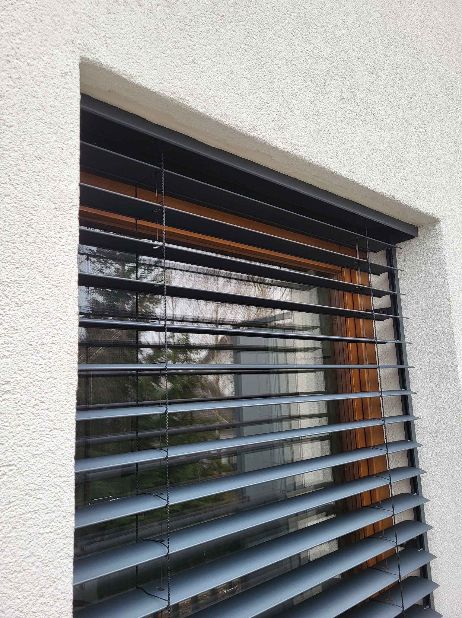 Szare żaluzje fasadowe zamontowane na oknie z drewnianą ramą, widok z zewnątrz na elewację budynku z fakturą baranka.