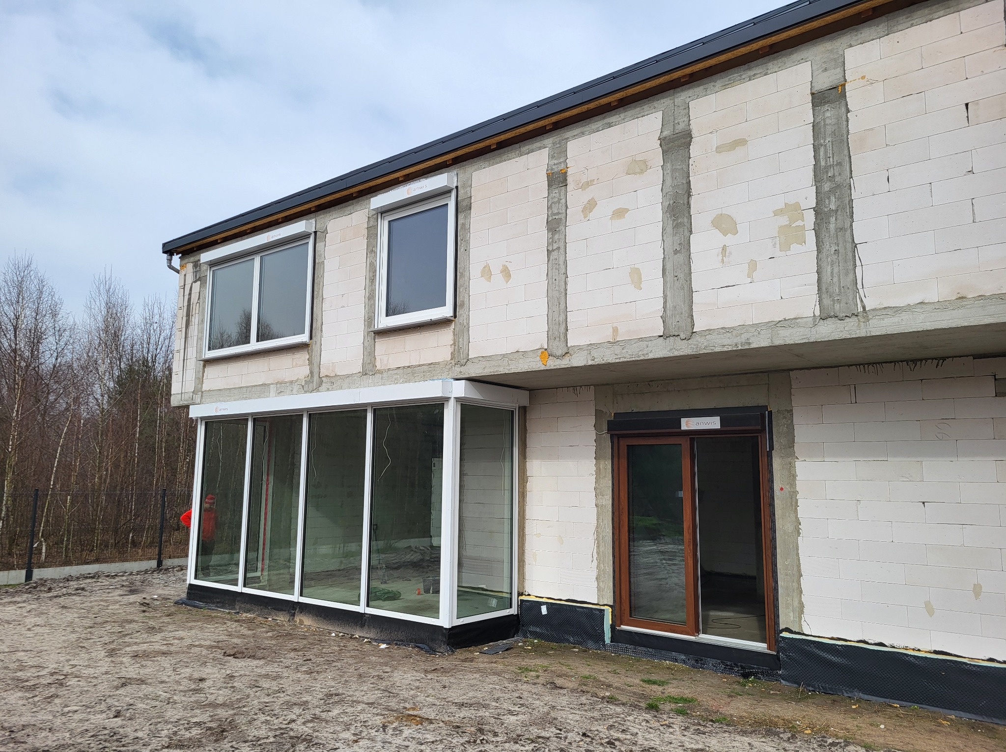 Niedokończony budynek z białego bloczka z zamontowaną przeszkloną werandą, oknami z roletami zewnętrznymi i brązowymi drzwiami przesuwnymi.