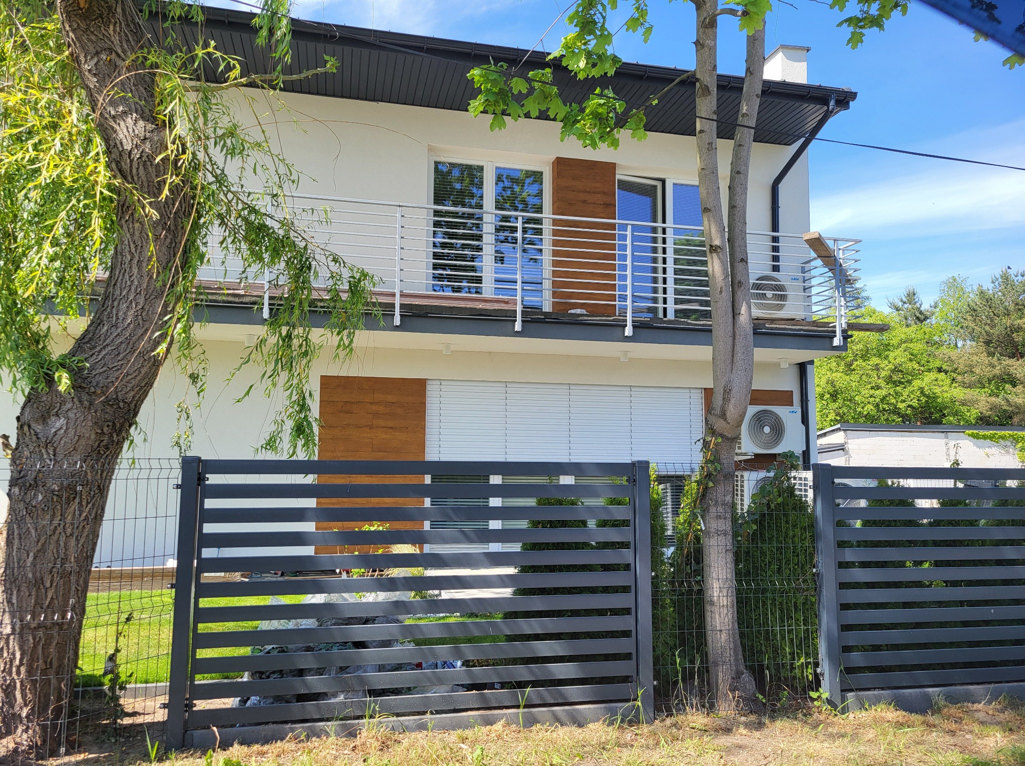 Dwukondygnacyjny dom z balkonem z metalową balustradą, widok zza grafitowego ogrodzenia panelowego, drzewa po bokach.