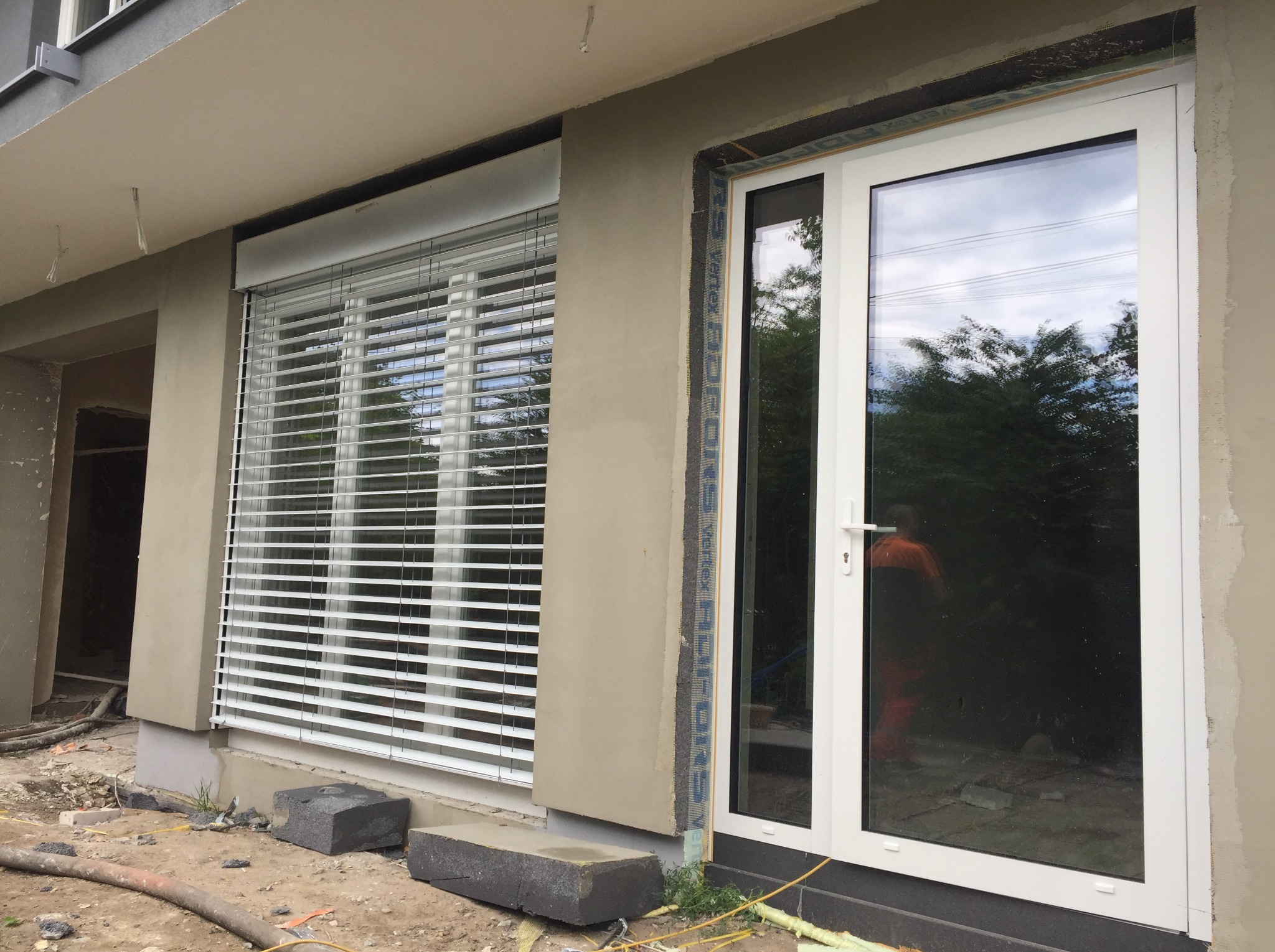 Białe okno z szybą refleksyjną i białe żaluzje poziome na elewacji budynku w trakcie wykończenia, widoczne przewody i gruz na ziemi.