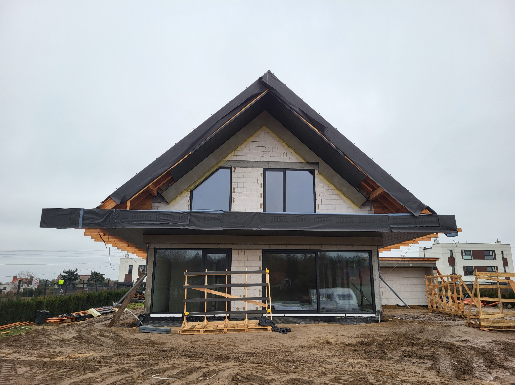 Dom w trakcie budowy, widoczne zamontowane okna z czarnymi ramami, ściany z białego betonu komórkowego, zabezpieczona membrana dachowa, teren wokół budynku rozkopany.