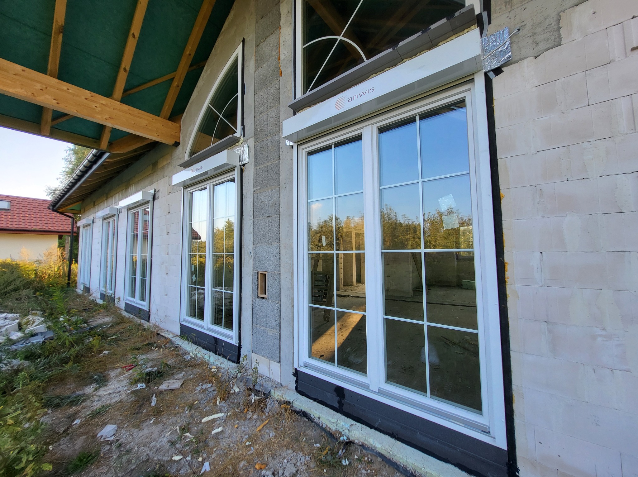 Białe okna z roletami zewnętrznymi w trakcie montażu w budynku z szarej cegły, widoczne materiały izolacyjne i logo firmy na rolecie.