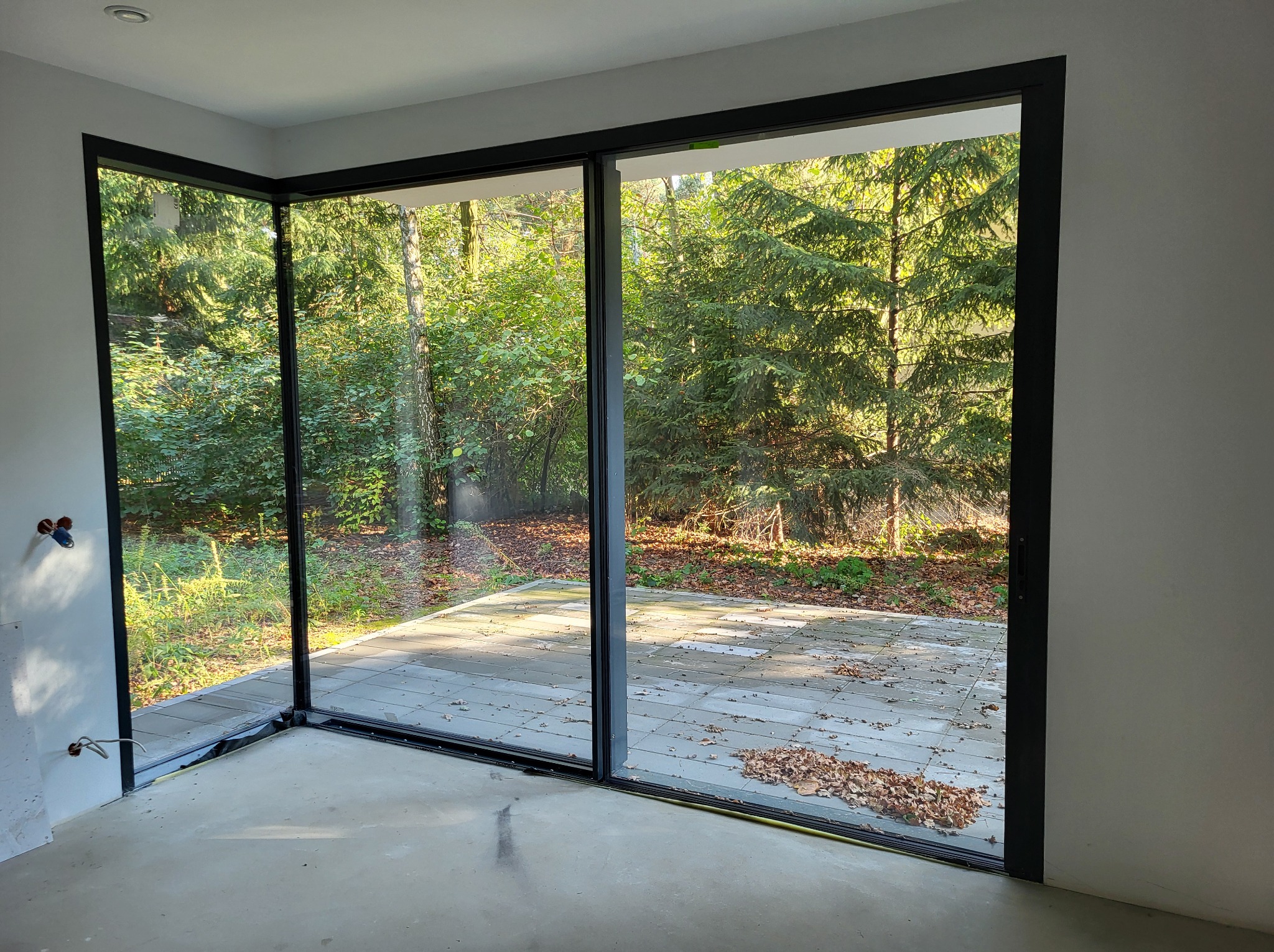 Nowoczesne, narożne okno przesuwne z czarnymi ramami, widok na zewnątrz z drzewami i tarasem, surowy stan wnętrza z widocznymi przewodami.