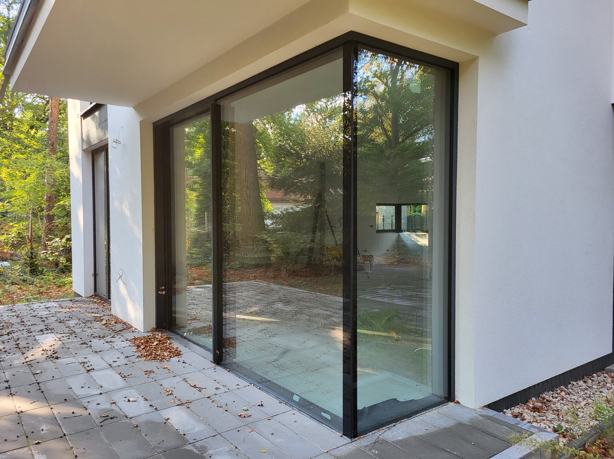 Nowoczesne, panoramiczne przeszklenie z czarnymi ramami w minimalistycznym budynku, widok na ogród, jesienne liście na tarasie z szarych płyt.