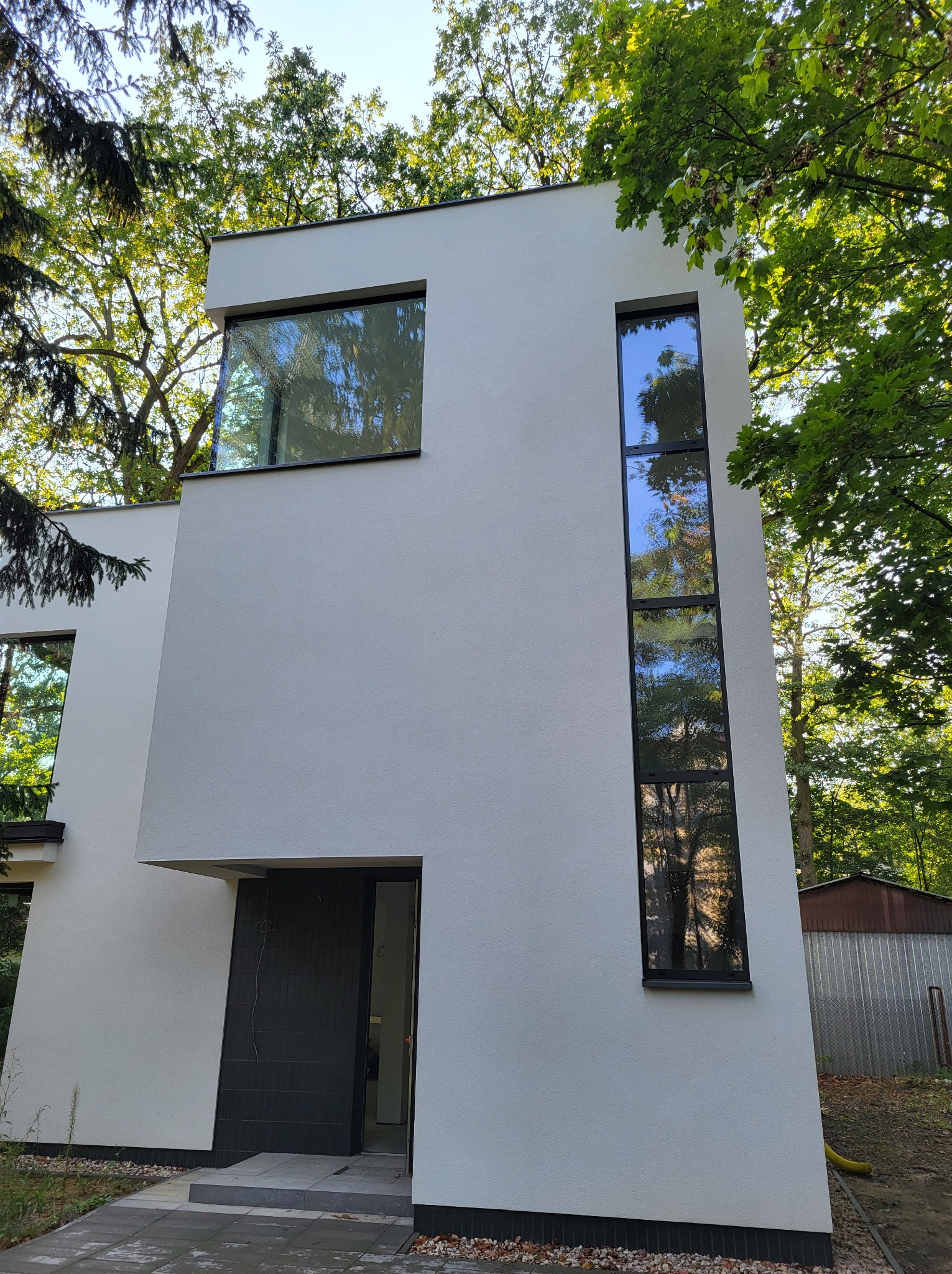 Nowoczesny dom z białymi ścianami, czarnymi ramami okiennymi i wysokim, wąskim oknem, otoczony zielenią drzew.
