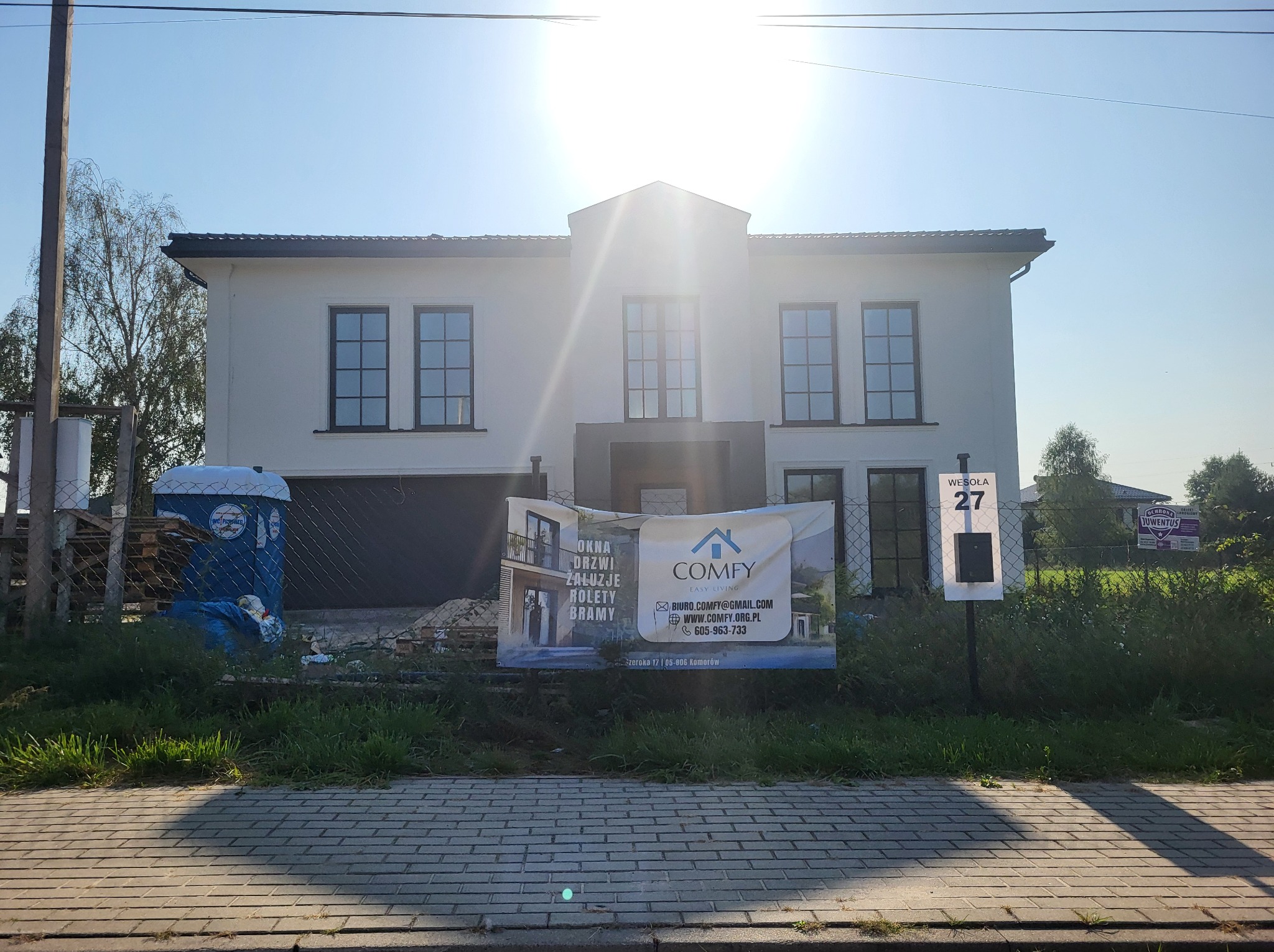 Nowy dom jednorodzinny z zamontowanymi oknami, widoczny baner reklamowy firmy Comfy oferującej okna, drzwi, żaluzje, rolety i bramy, na pierwszym planie chodnik z kostki brukowej i przenośna toaleta.