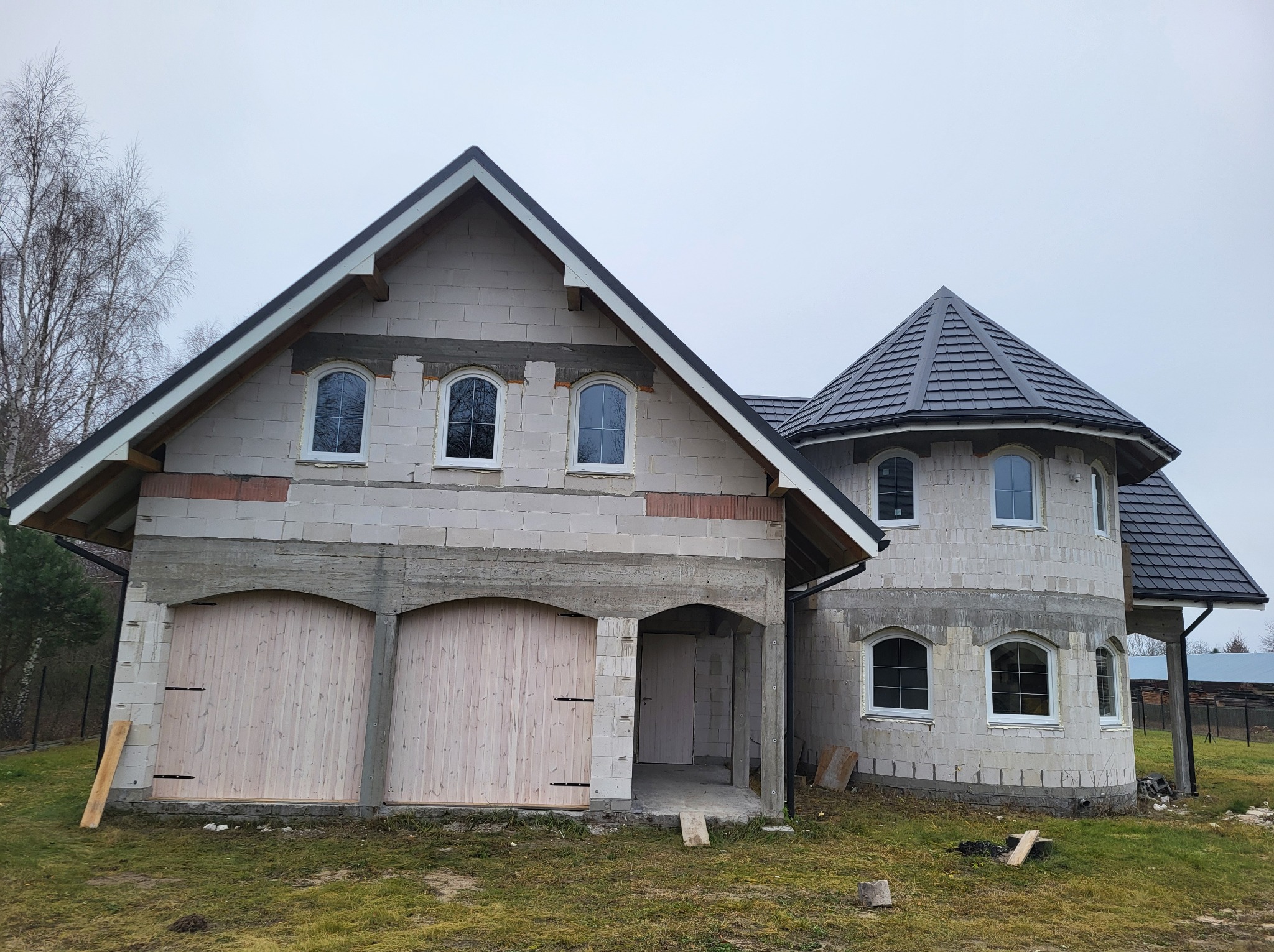 Surowy stan budynku w trakcie budowy z widocznymi białymi oknami z łukami, drewnianymi bramami garażowymi i szarą dachówką na dachu, otoczony jesienną trawą.