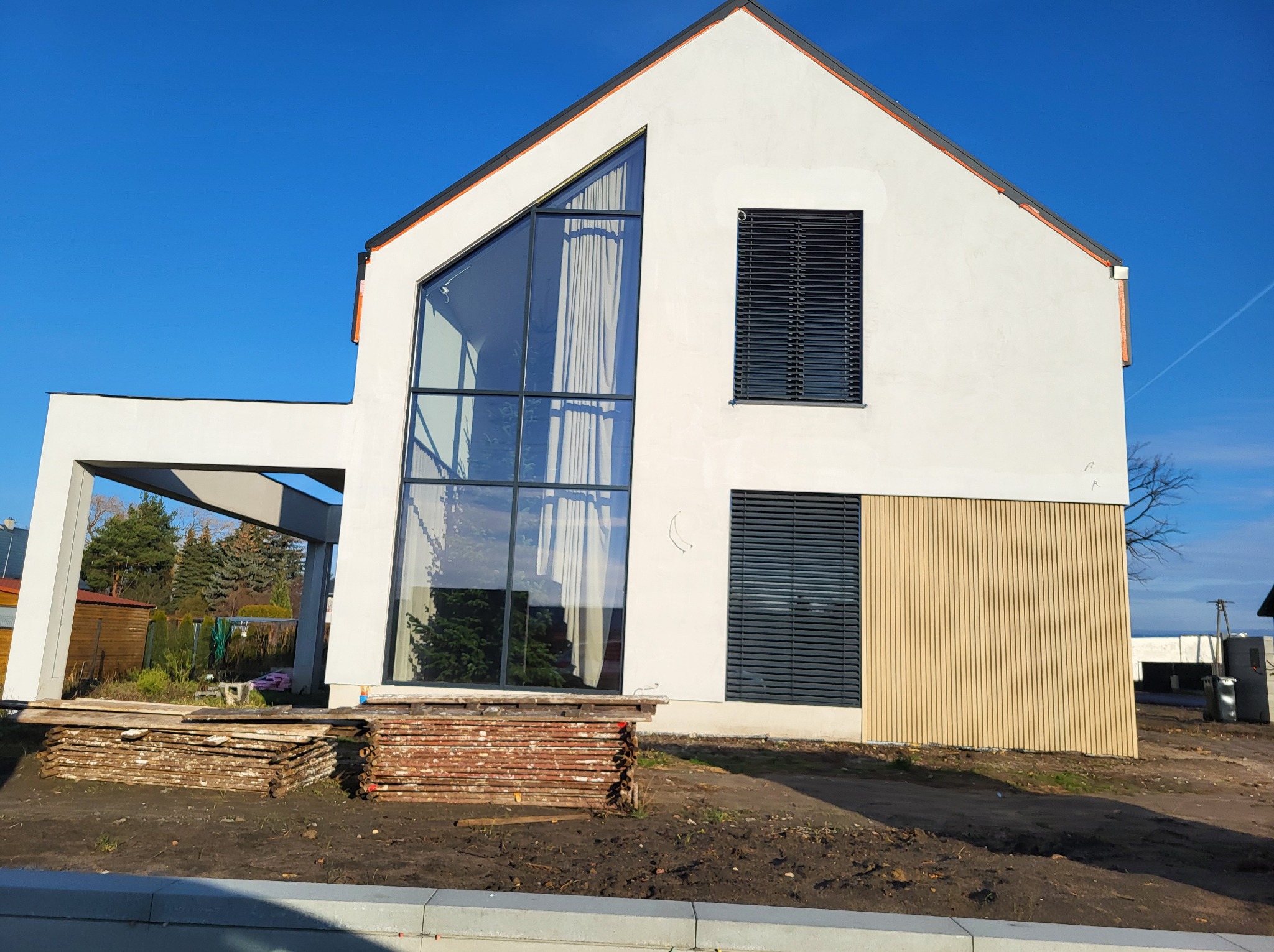 Nowoczesny dom z dużym trójkątnym oknem i ciemnymi żaluzjami poziomymi, widoczne drewniane panele elewacyjne i stosy desek na placu budowy