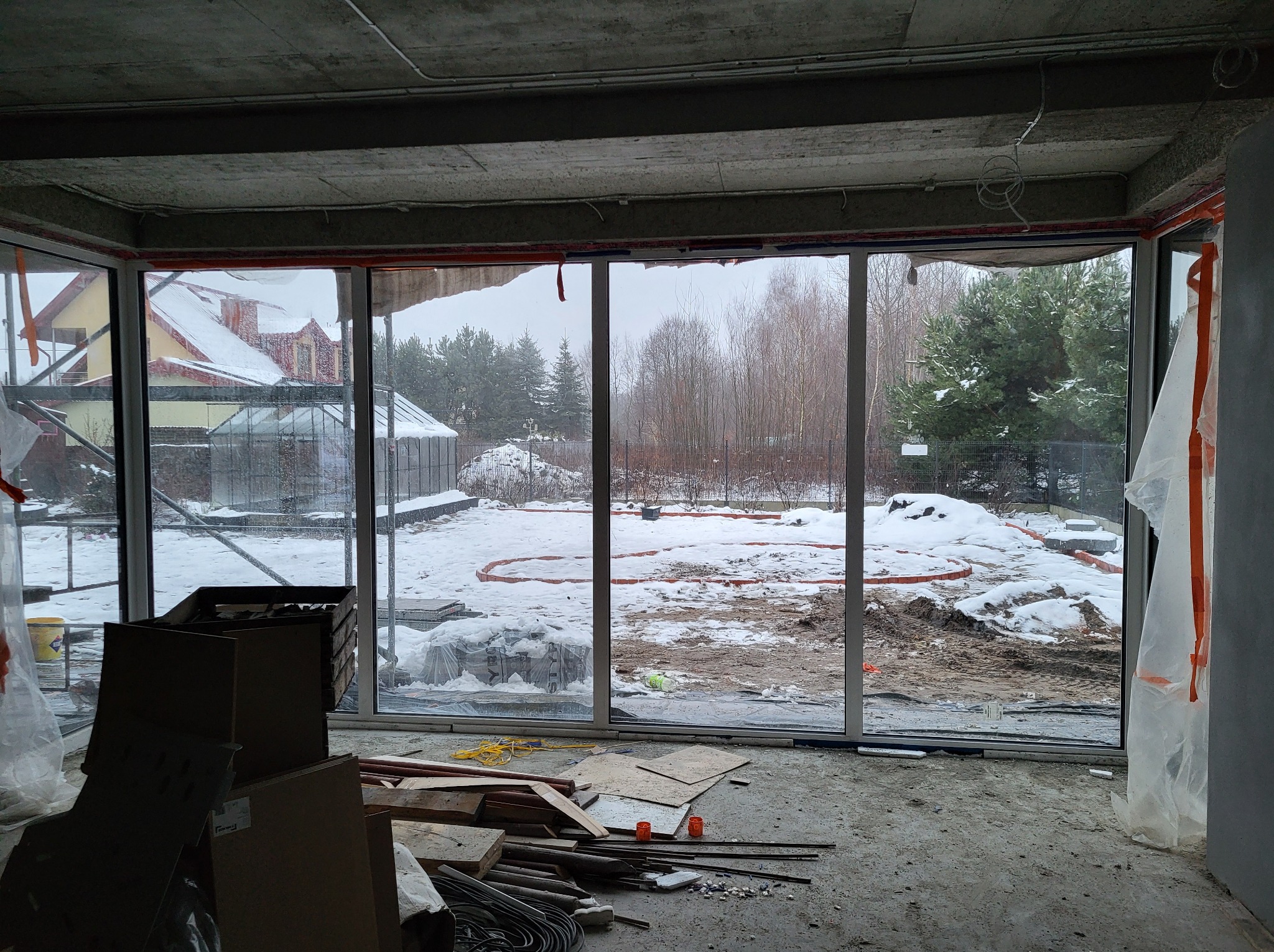 Wnętrze surowego domu w trakcie budowy z zamontowanymi dużymi przeszkleniami z widokiem na zasypany śniegiem ogród i sąsiedni dom