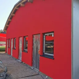 Czerwona elewacja budynku z drewnianymi elementami konstrukcyjnymi dachu, okna zabezpieczone folią, widoczne ślady prac budowlanych wokół budynku.