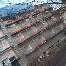 Budynek wielorodzinny w trakcie prac elewacyjnych, widoczne rusztowania, okna zabezpieczone folią, świeżo nałożony tynk, fragment nieba z nagimi gałęziami drzew