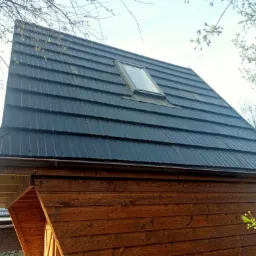 Ciemna, metalowa dachówka modułowa z oknem dachowym na drewnianej konstrukcji, widok z dołu, wiosenne gałęzie drzew w tle.