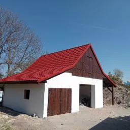 Nowo wybudowany, otynkowany i pomalowany na biało budynek gospodarczy z czerwonym dachem z blachodachówki i drewnianymi wrotami garażowymi, widoczne ślady prac wykończeniowych wokół.