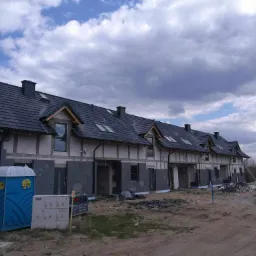 Szeregówka w trakcie budowy: surowe elewacje z widoczną izolacją, czarne dachy z dachówkami, niebieska toaleta przenośna na pierwszym planie, pochmurne niebo w tle.