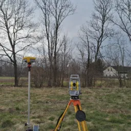 Dwa urządzenia pomiarowe: żółto-szary teodolit na statywie i odbiornik GNSS na tyczce, na tle łąki i drzew bez liści, w tle widoczny budynek.