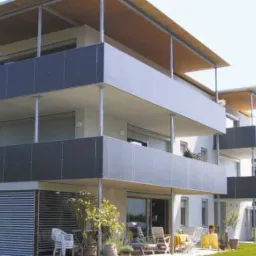 Nowoczesny budynek wielorodzinny z balkonami zabezpieczonymi szarymi panelami i zadaszeniem, widoczne meble ogrodowe na balkonie na parterze.