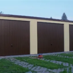 Trzy brązowe bramy garażowe segmentowe w jasnobeżowym budynku, widoczne na tle zielonego trawnika i drzew, z pomarańczowym przedłużaczem leżącym na trawie.