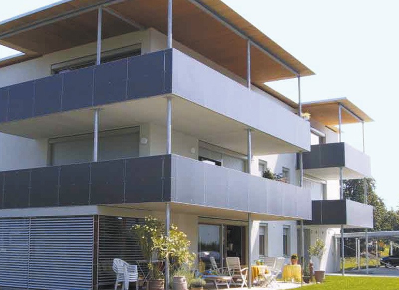 Nowoczesny budynek wielorodzinny z balkonami zabezpieczonymi szarymi panelami i zadaszeniem, widoczne meble ogrodowe na balkonie na parterze.