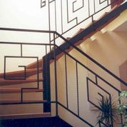 Klatka schodowa z metalową balustradą o geometrycznym wzorze, drewniane stopnie, kremowe ściany, roślina w doniczce u dołu schodów.