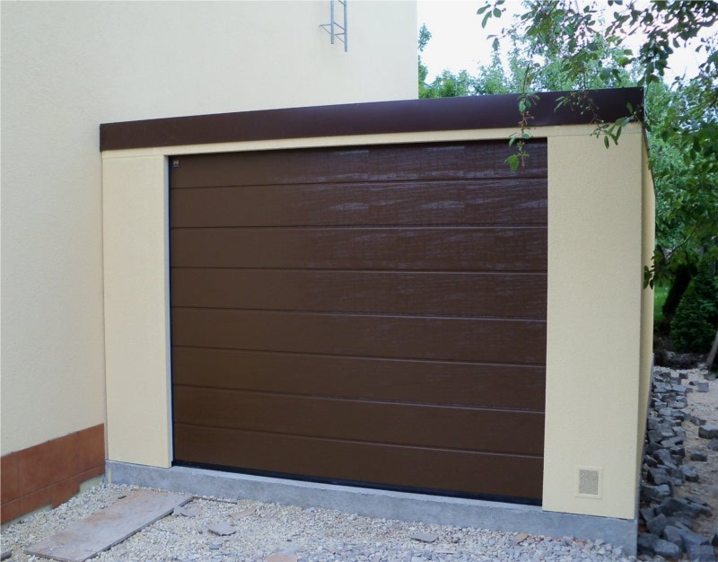 Zewnętrzna brama garażowa segmentowa, brązowa, z poziomymi panelami, osadzona w beżowej ramie budynku, widok z zewnątrz na tle zieleni.